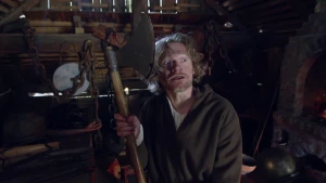 Een foto van een man die in een donkere, houten schuur staat waar bruine kettingen aan de muur hangen. De man kijkt met opengesperde ogen naar rechts. Hij heeft in handen een houten handvat met een metalen hakbijl eraan. Hij draagt een bruin kostuum.