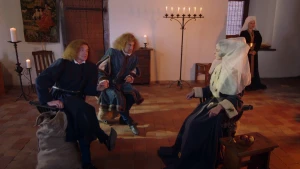 Een foto met daarop links twee mannen die op een stoel zitten met gekruisde benen. Ze kijken naar rechts waar een vrouw verkleed als Jacoba van Beieren zit in een donkerkleurig kostuum met witte mouwen en met een witte, doorzichtige sluier op haar hoofd. 