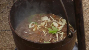 Een foto van een bruine ketel met een bruin handvat die aan een rechtvooraan deels zichtbare bruine paal hangt. Er zit een bruine smurrie in met enkele groene en witte blaadjes, gele maiskorrels en rode stukjes eten erin. Er stijgt witte damp uit op.