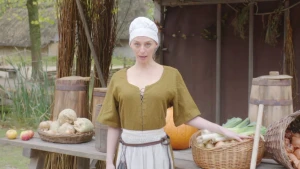 Een foto van een vrouw in een witte rok met bruine riem, een lichtbruine blouse en met een witte hoed of kap op. Haar hand ligt op een rieten mand met groente, uien en prei, erin op een tafel. Verder liggen er appels, een pompoen en koolrapen op.