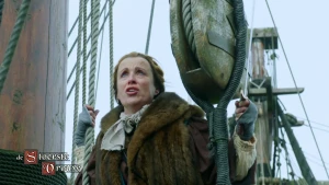 Een foto van een vrouw op een schip die een bruine bondmantel draagt en zich vasthoudt aan twee witte touwen. Links is een deel van een bruine paal te zien. Rechts hangt een katrol, een takelblok met een touw eraan. Linksonder staat "De stoerste vrouw".