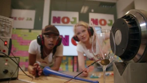 Een foto van Eva en Janouk achter een tafel met gehoorbeschermers en een veiligheidsbril. Links staan geluidsapparaten met druk- en draaiknopjes, rechts een luidspreker naast een wijnglas met plastic rietje erin en vastgeplakte rode en zwarte draden erop.