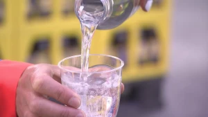 Een foto van een hand met een rode mouw die een doorzichtig glas vastheeft met daarin omhoog borrelende bubbels. Boven het glas wordt er doorzichtige frisdrank in het glas geschonken vanuit een doorzichtige fles.