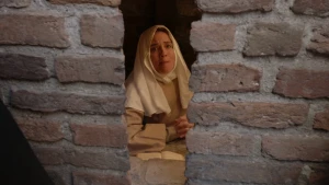 Een foto van een vrouw verkleed als Non Suster Bertken in lichtbruine kleding en witte doek om haar hoofd voor een tafel met een boek. Ze zit achter een spleet van een muur met roodbruine stenen. Ze zit met gevouwen handen te bidden en kijkt serieus. 