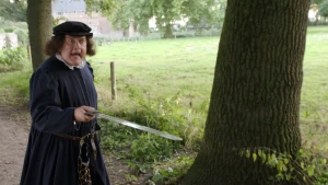 Een foto van een man verkleed als François Villon die met opengesperde ogen en open mond naar rechts kijkt. Hij heeft een zilverkleurig zwaard vast en wijst ermee naar rechts. De man draagt een donker pak en een pet. Hij staat op een pad naast een boom.