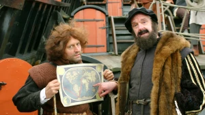 Een foto van twee mannen op een boot van zwart hout met rode deuren. De linkerman heeft een oude wereldkaart vast, de rechterman wijst ernaar. De rechterman is ontdekkingsreizer Magalhães in een bruine bondjas, zwarte pet en grijs pak met gele strepen.