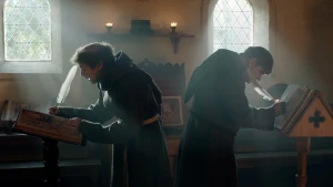 Een foto van twee mannen in een pei een bruin pak met capuchon. Ze staan met de rug naar elkaar met een veer in een boek te schrijven. De kamer is verlicht door twee ramen met zwarte driehoeken ervoor. Ze zijn monniken, die samen wonen en geloven in god.