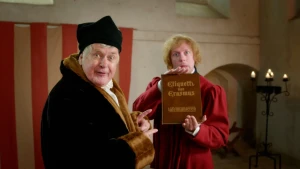 Een foto van twee verklede mannen. Eén is verkleed als Erasmus. Hij draagt een zwarte jas met bruine mouwen en kraag en een zwarte hoed. Hij wijst naar het boek dat een man in rood kostuum vastheeft waarop staat "Etiquette van Erasmus tafelmanieren".