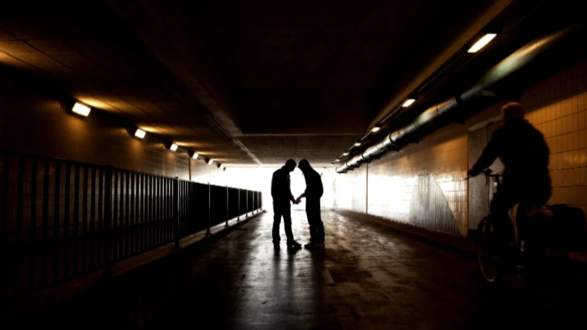 Een foto van twee mensen van veraf in een donkere tunnel met weinig licht. De ene mens geeft de andere iets aan in zijn of haar hand, maar wat is door het gebrek aan licht niet te zien. Rechts is een fietser te zien die langs de twee mensen zal rijden.