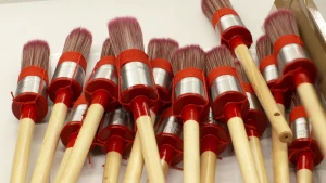 Een foto met zestien kwasten erop. Het zijn verfkwasten om muren of andere dingen mee te verven. De kwasten hebben een lichtbruin houten handvat dat aan een zilverkleurig deel met rode stof eromheen zit. De lange, rechtopstaande haartjes zitten bovenop.