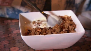 Een foto van een wit schaaltje met granola en yoghurt erin op een bruine tafel met cirkelvormen erop. Een zilverkleurige lepel roert erdoorheen. De granola is een mengsel van bruine granen, rozijnen, pitjes en niet-zichtbare stukjes appel en kaneel.
