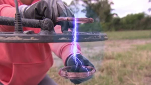 Een foto van het zwarte, ronde blad van een metaaldetector en de stekker die er bovenop uitsteekt. Iemand houdt twee spoelen rood metaalraad boven en onder het blad. Daartussen is een paars met wit magnetisch veld te zien, dat lijkt op een bliksemflits.