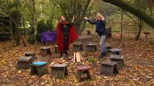 Een foto van een bos. Janouk en Margarita, de heks van Appelscha, staan in een kring van dertien houten krukjes voor een klein vuur met houten planken. Ze houden hun handen met hun handpalmen omhoog. Margarita draagt een zwart pak met rode cape.