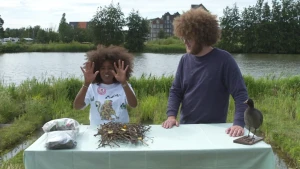 Een foto van Eva en Florian op een groen grasveld aan de waterkant. Op de tafel ervoor ligt een stapel takken en twee plastic zakken met dingen erin. Rechts staat een opgezette vogel, een meerkoet, zwart met witte snavel. Eva houdt beide handen omhoog.  