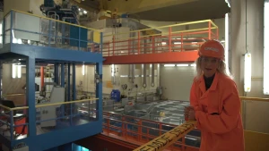 Een foto van Janouk die in een kerncentrale staat. In de centrale wordt energie, stroom opgewekt. Janouk draagt een oranje pak en een oranje plastic helm met het woord "bezoeker" erop.  Achter Janouk liggen witte platen op een grijze vloer.