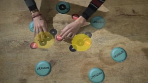 Een foto van een houten vloer met zes groenblauwe rondjes met het woord 'Uranium", zes rode rondjes met het woord "Afval" en zes zwarte rondjes met de letter "N", voor Neutron. Om twee groepjes rode en zwarte rondjes staat een doorzichtig, geel rondje.