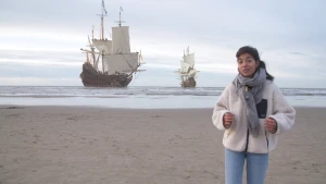 Een foto van een presentator op het strand. In het water zijn twee op de foto toegevoegde Nederlandse handelsschepen te zien. De schepen zijn van hout en hebben meerdere witte zeilen en Nederlandse vlaggen in de masten. 