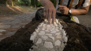 Een foto van een voorbeeld van een Romeinse weg tussen twee stroken aarde. Zo'n weg bestaat uit kleine, witte steentjes met een dikke laag zand erop en tussen het zand witte, Romeinse stenen. De presentator, gezicht niet te zien, drukt een steen erin.