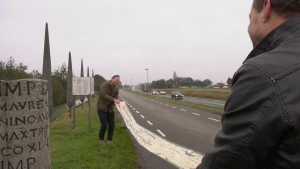 Een foto van twee mannen op een grasveld langs een autoweg. Ze zijn bezig een lange Romeinse wegenkaart uit te rollen. Aan de linkerkant staat een grijze mijlpaal, een ronde paal met de afstand naar en naam van de toekomstige plaats erop.