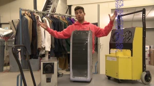 Een foto van Nizar achter een tafel met drie apparaten die  brandlucht kunnen verwijderen met draaiknoppen en drukknoppen. De meest rechtse is geel en heeft twee ronde cirkels met luchtgaatjes en een ronde pijp waar geanimeerde blauwe balletjes uitkomen.