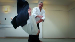 Een foto van twee mannen die de zelfverdedigingssport aikido  aan het doen zijn in een witte jas en blauwgrijze broek. Eén man heeft met één hand de rechterpols van de andere man vast. De andere man wordt omvergegooid en hangt met zijn voeten in de lucht.