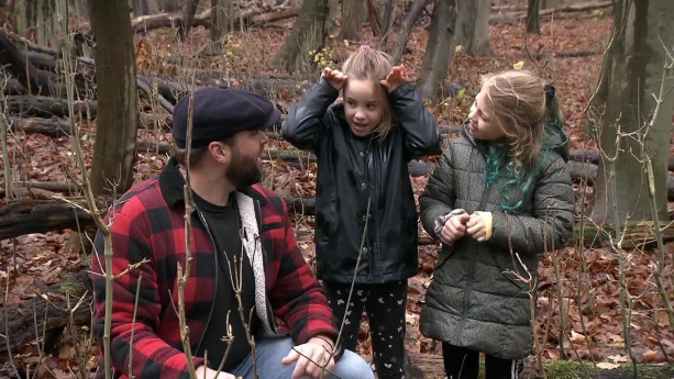 Een foto van presentator Tim die in het bos op de grond zit en met twee meisjes aan het praten is. Een van de meisjes houdt haar handen naast haar hoofd alsof ze een hert nadoet.