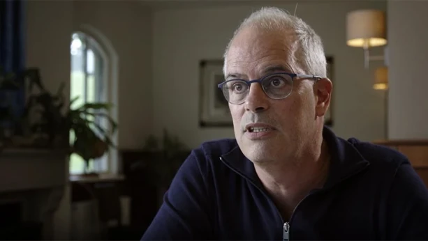 Een foto van een man in een kamer. Hij heeft grijs haar en een  bril op.  Hij kijkt naar iets naast de camera, en is wat aan het vertellen.