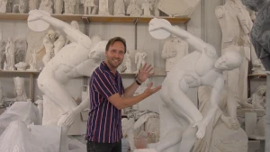 Een foto van een presentator in een kamer met witte beelden die met zijn handen wijst naar een wit marmeren beeld. Het beeld is van een naakte man die in één naar achteren gestrekte arm een discus houdt, een ronde schijf om mee te gooien bij discuswerpen.