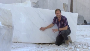 Een foto van een presentator die op zijn knie zit en zijn rechterhand heeft liggen op een groot, rechthoekig blok marmer dat wit is, met grijze kronkelige lijnen in het wit. Marmer is een steensoort ontstaan in de Italiaanse bergen uit kalk. 