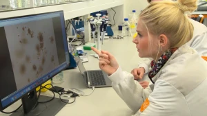 Een foto van Janouk in een witte laboratoriumjas en iemand in dezelfde jas, waarvan het gezicht niet te zien is. Ze wijst naar een beeldscherm op tafel, met bruine, ronde vlekken en vlekjes erop. Dat zijn tumorcellen van mensen met de ziekte kanker.