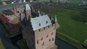 Een foto van een kasteel van steen met een gracht van water eromheen. Het heeft vier torens met een grijs puntdak, één schuin dak, één toren met een schuin dak en ramen met roodwitte luiken. Achteraan zijn de binnenplaats en poort van het kasteel te zien.
