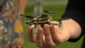 Een foto van een hand die een kleine drone vastheeft. De onderkant is van doorzichtig plastic, met aan vier kanten een zwarte propeller met twee wieken. Bovenop zitten computerchips op een plaat, rode en gele stroomdraden en een geheugenkaart.