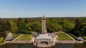 Een foto van een stenen gebouw, Jachthuis Sint Hubertus. Vooraan ligt water achter een muur met aan beide kanten ronde, grijze daken. Het gebouw heeft een grijs dak, een grote toren met schuin dak achteraan en een tuin van groen gras aan beide kanten. 