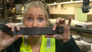 Een foto van Janouk die een geel veiligheidsvest met zilverkleurige strepen over haar kleding aanheeft in een fabriek. Ze houdt een langwerpig stuk zwart rubber voor de camera, dat gebruikt wordt voor het maken van een autoband. 