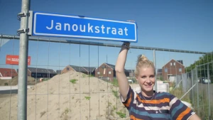 Een foto van Janouk die lachend naar de camera kijkt. Ze heeft met haar rechterhand een blauw straatnaambord vast boven haar hoofd met de straatnaam "Janoukstraat" erop. Het bord zit aan een stalen paal vast. Erachter staan hekken voor een berg zand.