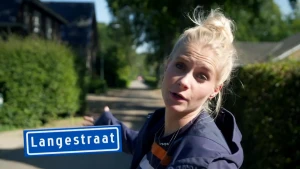 Een foto van Janouk in het midden van een straat die naar achteren wijst met haar rechterhand. Ze staat op een lang pad met aan beide kanten  groene struiken en huizen. Naast Janouk staat een blauw straatnaambord met de straatnaam "Langestraat" erop.