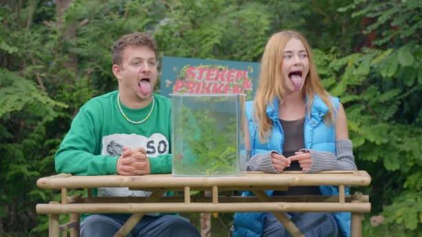 Een foto van presentatoren Emma en Jurre die buiten aan een tafel zitten met uitgestoken tongen. Op de tafel staat een bak waar je doorheen kan kijken waar hommels en planten inzitten.