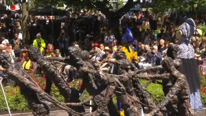 Een foto van een beeld van acht mensen die voorovergebogen lopen en vastzitten aan stalen kettingen. Daarachter is de rug van twee mensen in blauwgele en witblauwe kostuums te zien en zitten mensen die Keti Koti, afschaffing van de slavernij, vieren.