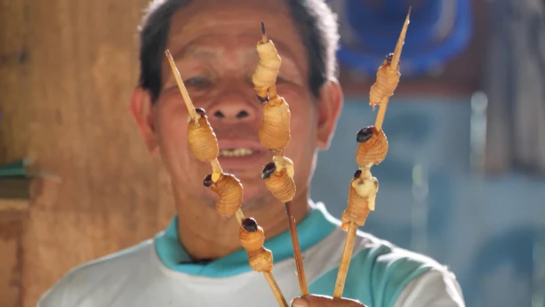 Een foto van een man in Peru. Hij houdt drie stokjes met gegrilde wormen voor de camera. 