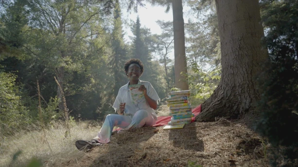 Een foto van presentator Tirsa. Ze zit op de grond in een bos, naast een grote boom. Naast haar staat een stapel boeken. Tirsa kijkt vrolijk in de camera.