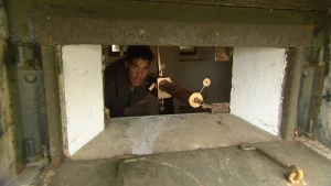 Een foto van de presentator met een automatisch geweer van vroeger. Hij is te zien door een gat in een dikke betonnen muur. Voor het gat ligt een metalen plaat met haken.