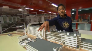 Een foto van de presentator achter een model van de Van Nellefabriek in Rotterdam. Er is een lang gebouw met delen in verschillende hoogte. De presentator wijst hier overheen naar een gebouw dat er voor staat. Achter haar staan meer modellen van gebouwen.