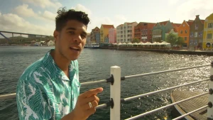 Een foto van de presentator in Willemstad, de hoofdstad van Curaçao. Hij staat bij een brugleuning en achter hem is een rivier en opvallend gekleurde huizen.