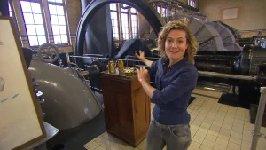 Een foto van de presentator Nienke in een ruimte met grote machines en buizen. Ze wijst naar achteren, naar een groot wiel dat met stangen en buizen vastzit aan de rest. Achter het wiel staan even grote wielvormige kasten.