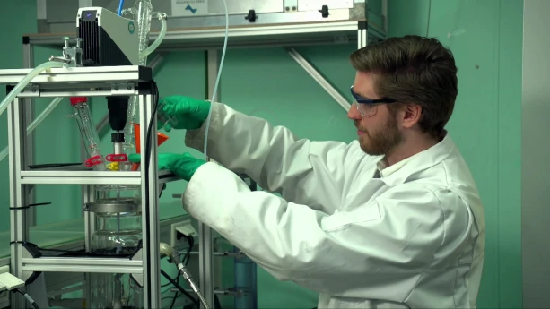 Een foto van iemand in een witte jas met een veiligheidsbril op en groene handschoenen aan. Hij staat bij een metalen rek met een glazen pot en plastic slangetjes. Hij doet iets in de pot met een trechter.