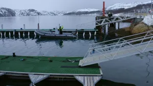 Een foto van twee pieren met loopplanken in een haven in Noorwegen. Bij de achterste pier ligt een rubberbootje met iemand daarin. Op de achtergrond zie je besneeuwde bergen.