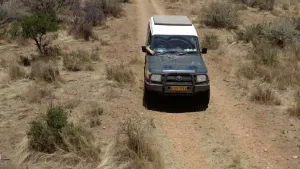 Een foto van een auto met vierwielaandrijving. Hij staat op een onverharde weg. Je ziet dit beeld vanuit de lucht. Een persoon in de auto houdt een antenne vast, die op de motorkap ligt.