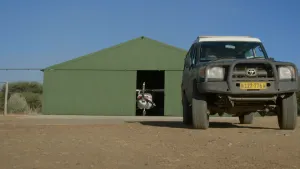 Een foto van een auto met vierwielaandrijving. Hij staat op een onverharde weg. Op de achtergrond zie je een opslagplaats voor een of meer vliegtuigen. De deur daarvan staat open en je ziet een propellervliegtuig staan.