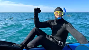 Een foto van bioloog Freek in een duikpak. Hij is op zee en zit op de rand van een rubberboot en steekt zijn vuist omhoog. Op de achtergrond zie je dolfijnen zwemmen.