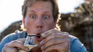 Een foto van bioloog Freek. Voor de camera duwt hij met een takje de bek open van een slang. Je ziet twee grote giftanden in de bek. Freek kijkt er met open mond naar.